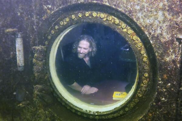 In this photo provided by the Florida Keys News Bureau, diving explorer and medical researcher Dr. Joseph Dituri peers out of a large porthole, Saturday, May 13, 2023, at Jules' Undersea Lodge positioned at the bottom of a 30-foot-deep lagoon in Key Largo, Fla. On Saturday, Dituri broke a record for the longest time living underwater at ambient pressure, his 74th day of a planned 100-day mission living submerged in the Florida Keys. The previous underwater habitation record of 73 days, two hours and 34 minutes was set by two professors from Tennessee in 2014 at the same location. Dituri's underwater mission, dubbed Project Neptune 100, was organized by the Marine Resources Development Foundation and combines medical and ocean research along with educational outreach. (Frazier Nivens/Florida Keys News Bureau via AP)