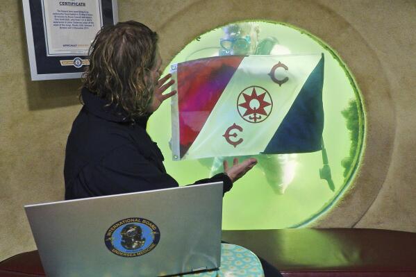 In this photo provided by the Florida Keys News Bureau, Dr. Joseph Dituri, left, peers out a large porthole from inside Jules' Undersea Lodge, located 30 feet below the surface in a lagoon Wednesday, March 1, 2023, in Key Largo, Fla., to see diver and Jules' founder Ian Koblick delivering a prestigious Explorer's Club flag. Dituri submerged Wednesday to begin a planned 100-day dive in the habitat, hoping to establish a new record for underwater human habitation at ambient pressure while educating young divers and conducting medical and marine science research. (Frazier Nivens/Florida Keys News Bureau via AP)